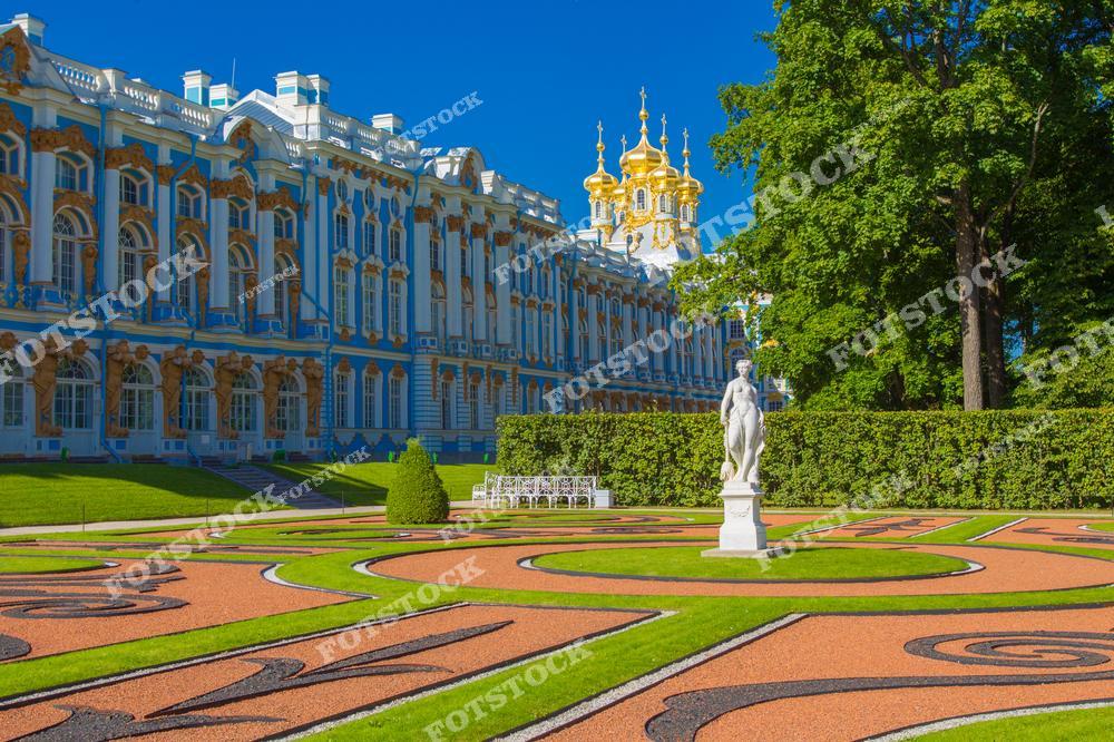 На фотографии изображен Екатерининский дворец в Царском Селе, одно из самых известных произведений барочной архитектуры в России. Великолепные золотые купола и богатые фасады здания создают атмосферу роскоши. На переднем плане находится декоративный сад с ухоженными газонами и изящной статуей, добавляющей гармонии общему виду. Это место является популярной достопримечательностью, привлекающей туристов своим великолепием и историческим значением.